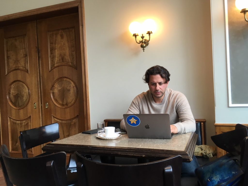 Ein Mann sitzt an seinem Computer und schreibt konzentriert. Vor ihm steht eine Tasse mit Kaffee und auf der Bank neben ihm liegt eine Tasche